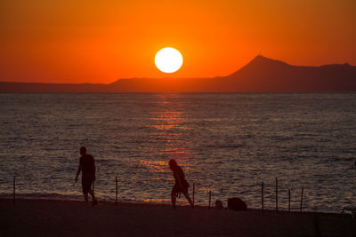 Silhouette man and woman walking at beach against sky during sunset
