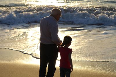 Rear view of man with granddaughter standing on shore at beach during sunset