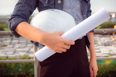 Midsection of architect holding blueprint and hardhat while standing outdoors