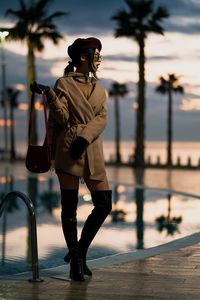 Full length of young woman standing at poolside against sky during sunset