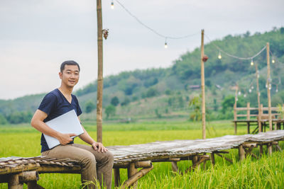 Portrait of man sitting with laptop on boardwalk at rice paddy