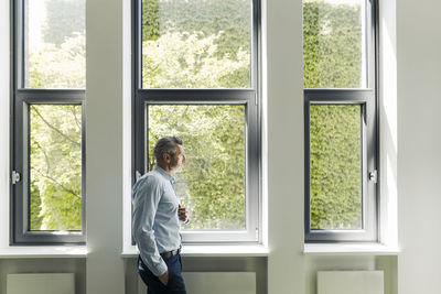 Businessman with hands in pockets looking through window while standing at office