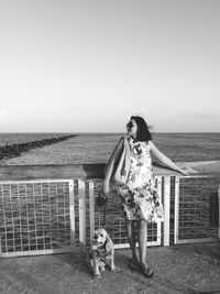 Full length of woman with dog standing by railing against sea and clear sky