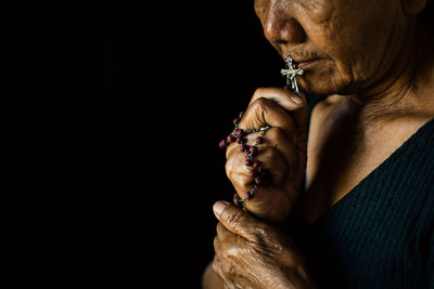 Close-up of senior woman holding rosary while praying against black background