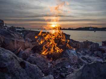 Close-up of campfire on rocks at lakeshore against cloudy sky during sunset