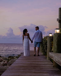Rear view of couple standing on shore against sky during sunset