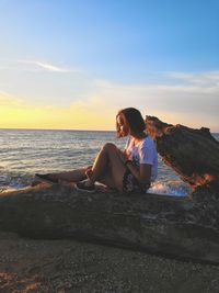 Side view of young woman sitting on driftwood at beach against sky during sunset