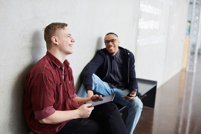 Smiling student holding digital tablet while sitting with friend on bench in university