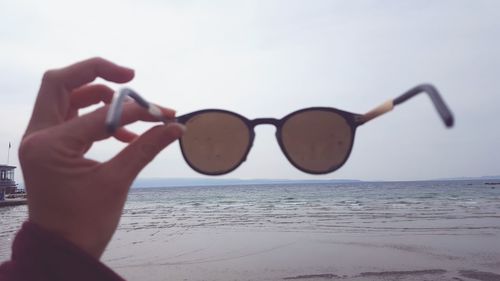 Midsection of person holding sunglasses at beach against sky