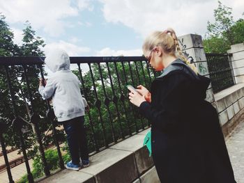 Rear view of girl by railing with woman using mobile phone while standing on footpath against sky