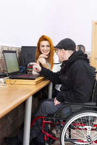 Smiling female caregiver talking with disabled man by laptop at nursing home