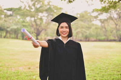 Portrait of young woman in graduation gown holding certificate while standing at park