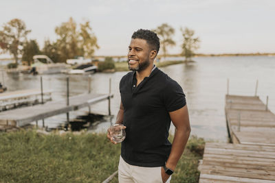 Smiling man standing with wineglass and hand in pocket by pier