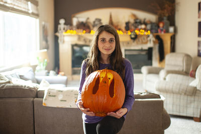 Tween girl 10-12 years old holds up carved pumpkin in living room