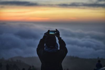 Woman photographing at camera against sky during sunset