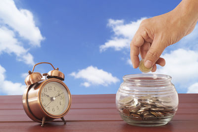 Close-up of hand putting coin in jar with alarm clock on table