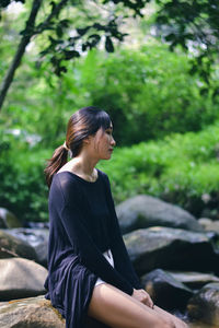 Side view of young woman looking away while sitting against trees