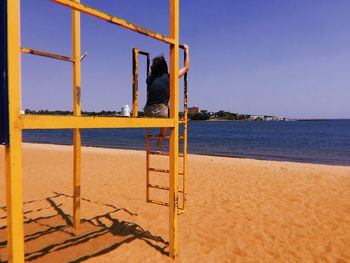 Rear view of young woman sitting by beach against sky