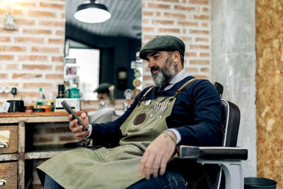 Side view of middle aged male barber sitting on chair in barbershop and browsing mobile phone while having break during work