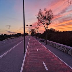 Empty road by trees against sky during sunset