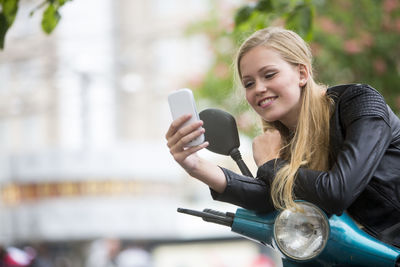 Happy woman using phone while leaning on motor scooter handle