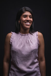 Positive self assured young latino female wearing casual blouse smiling and looking away while standing against black background