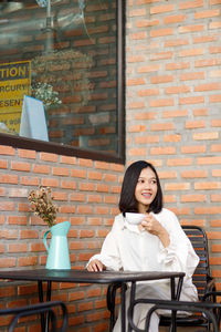 Young beautiful asian woman in white blazer with coffee cup sitting in cafe dark brown background