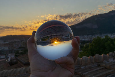 Close-up of hand holding crystal ball against sky during sunset