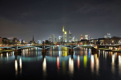 Illuminated bridge over river by buildings against sky at night