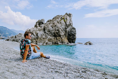 Man sitting on rock at beach against sky