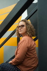 Young adult millennial with curly hair sits on steps. student looks away. lifestyle