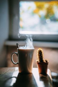 Close-up of coffee cup by cactus on table against window at home