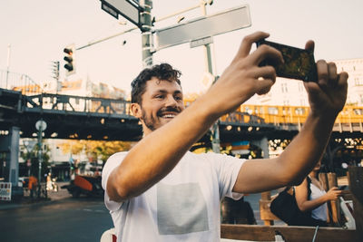 Smiling young man photographing through smart phone while standing on street in city