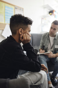 Depressed boy sitting with head in hand by male counselor discussing at school office