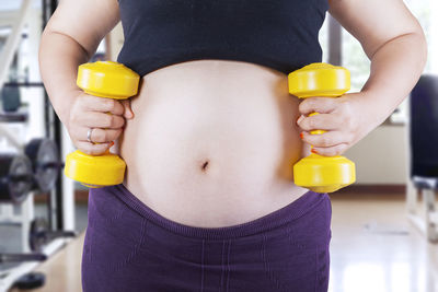 Midsection of pregnant woman holding yellow dumbbells in gym