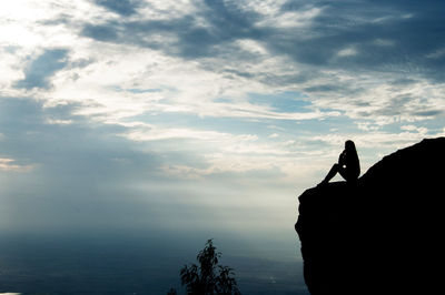 Silhouette woman sitting at the edge of cliff against cloudy sky