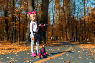 Full length portrait of smiling girl with push scooter standing against trees during autumn