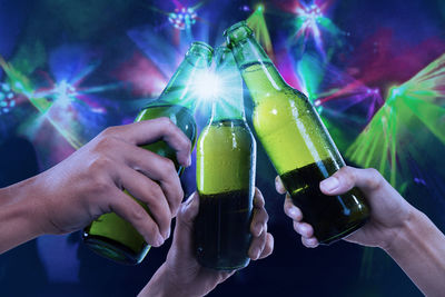 Close-up of hands toasting beer bottles against illuminated lights