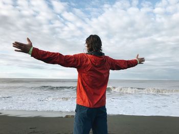 Rear view of man with arms outstretched while standing on beach against sky