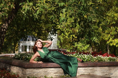 Portrait of woman sitting by tree against autumn park background, wearing green silk dress 