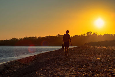 Rear view of man standing on shore against sky during sunset