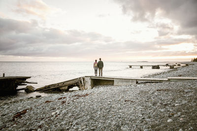 Rear view of couple standing on jetty at beach against sky during sunset