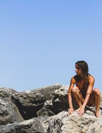 Young woman crouching on rock against clear blue sky