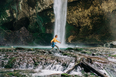 A young active woman in a yellow longsleeve walks through a mountain stream near a waterfall.