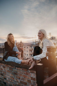 Cheerful women talking while sitting on building terrace against sky