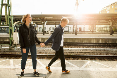 Full length of young man pulling teenage girl on skateboard at railroad station platform