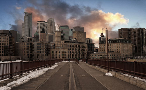 Road leading towards buildings in city against cloudy sky