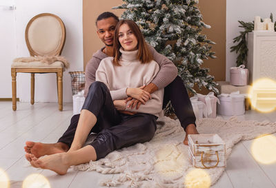 A happy stylish guy in a brown sweatshirt is sitting hugging his girlfriend, near the christmas tree