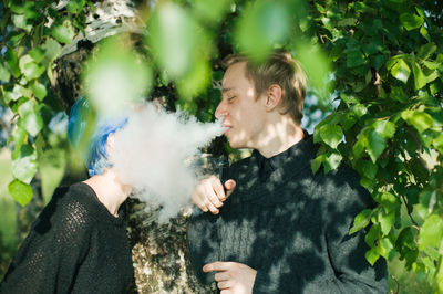 Young man exhaling smoke on friend by tree