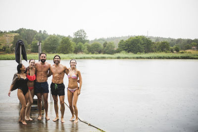 Full length portrait of happy male and female friends standing on jetty over lake during weekend getaway